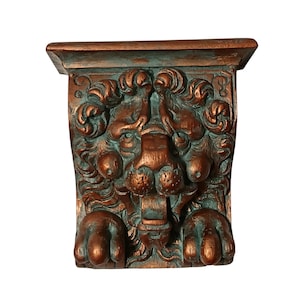 May include: A decorative wall corbel featuring a sculpted lion head with a prominent mane and two front paws at the base. The piece has a metallic copper finish with dark teal green oxidation accents in the recessed areas of the carving.
