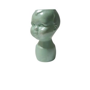 Könnte beinhalten: Ein blass mintgrüner Keramikübertopf in Form eines stilisierten Babykopfes. Das Objekt hat eine glatte, glänzende Oberfläche mit geformten Gesichtsdetails wie Augen, Nase und Lippen sowie einer offenen Oberseite für kleine Pflanzen oder Gegenstände.