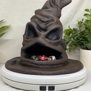 May include: A brown, textured, conical hat with a wide brim and a deep, open mouth-like cavity containing a collection of multi-colored, multi-sided gaming dice. The hat sits atop a white, circular, rotating display stand with two black switches.