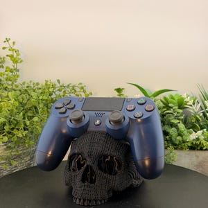 May include: A dark blue wireless game controller resting on top of a black, textured, skull-shaped stand. The controller features a central touchpad, two analog sticks, a directional pad, and four action buttons with triangle, circle, cross, and square symbols.
