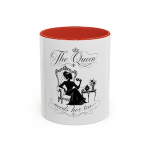 May include: A white ceramic mug with a bright red interior and rim. The front features a black silhouette graphic of a person in a gown sitting in a chair with a tea cup, accompanied by the text The Queen needs her tea and a small crown.