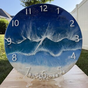 May include: A round wall clock featuring a resin ocean scene with deep blue water transitioning to light blue and white sandy shores. The clock face includes raised white numbers from 1 to 12, white hands, and a collection of small seashells at the bottom.