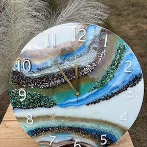 May include: A round wall clock featuring a multi-layered resin design with swirling patterns of white, blue, brown, and green. The clock face includes embedded crushed glass textures, white raised numerals, and two thin gold-colored metal hands.