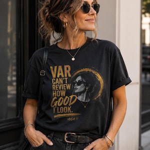 Peut inclure: Un t-shirt noir à manches courtes avec une impression graphique dorée représentant une personne portant des lunettes de soleil dans un cercle. Le texte indique : VAR CAN'T REVIEW HOW GOOD I LOOK. MUDA avec un cœur est imprimé en dessous.