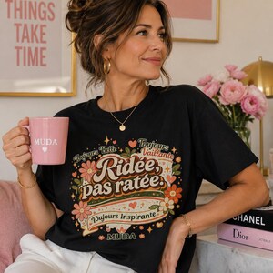 Peut inclure: Un t-shirt noir à manches courtes avec un imprimé graphique coloré portant le texte "Ridée, pas ratée" entouré de motifs floraux, d'étoiles et des phrases "Toujours Belle", "Toujours Vaillante" et "Toujours Inspirante" avec "MUDA" en bas.