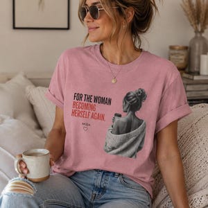 Peut inclure: Un t-shirt rose poudré avec un motif en noir et blanc représentant une personne en chignon tenant une tasse, accompagné du texte "FOR THE WOMAN BECOMING HERSELF AGAIN." et "MUDA" avec un petit cœur imprimé en noir et rouge sur le devant.