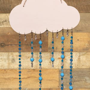 May include: A light pink wooden cloud cutout hanging from a jute twine string. Attached to the bottom edge are several dangling strands of blue and clear glass beads in varying sizes and shapes, creating a rain-like effect from the cloud.
