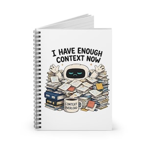 May include: A white spiral-bound notebook featuring a cartoon robot buried under a large pile of papers. The text above the robot reads I HAVE ENOUGH CONTEXT NOW, and a coffee mug in the foreground is labeled CONTEXT OVERLOAD in black text.