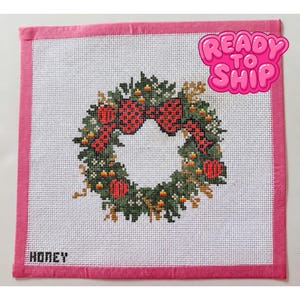 Könnte beinhalten: Ein quadratisches Stickbild mit einem Weihnachtskranz-Motiv aus grünem Laub, roten Ornamenten und Zuckermais-Akzenten. Die Leinwand hat einen rosa Stoffrand und das Wort HONEY in der unteren linken Ecke sowie eine READY TO SHIP-Grafik.