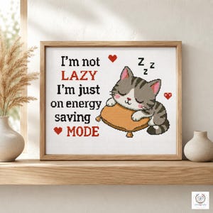 May include: A cross-stitch pattern in a wooden frame featuring a gray tabby cat sleeping on a tan pillow. The text reads: I'm not LAZY I'm just on energy saving MODE. Red hearts and black Zzz symbols surround the cat on a white fabric background.