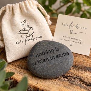 May include: A smooth dark gray stone engraved with the text nothing is written in stone, accompanied by a cream-colored drawstring pouch featuring a box and heart graphic with the text this finds you, and a card reading this finds you a little reminder for when you need it.