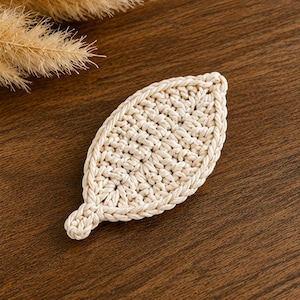 May include: A cream-colored crochet leaf ornament crafted from thick cotton cord. The leaf features a central vein pattern with a textured, knotted stitch design and a small stem at the base, showcasing intricate hand-stitched fiber art details.
