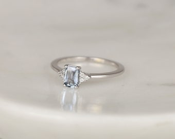 May include: A silver ring with a light blue gemstone in an emerald cut setting. The gemstone is surrounded by smaller, clear gemstones.