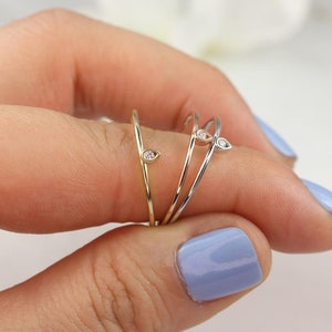 May include: Three delicate gold rings with a single small diamond on each. The rings are stacked on a finger.