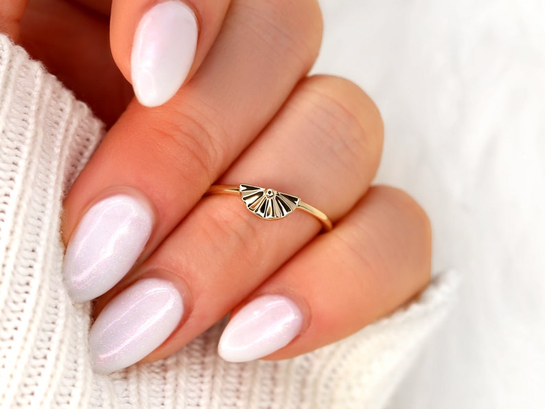 May include: A gold ring with a fan-shaped design. The ring is on a finger with white nail polish.