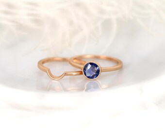 May include: Two rose gold rings. One ring features a round, bezel-set, blue gemstone. The second ring has a curved design. The rings are displayed on a white surface with soft, blurred background elements.