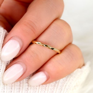 May include: A thin gold band ring with a hammered texture. The ring is worn on a finger with a light pink nail polish.