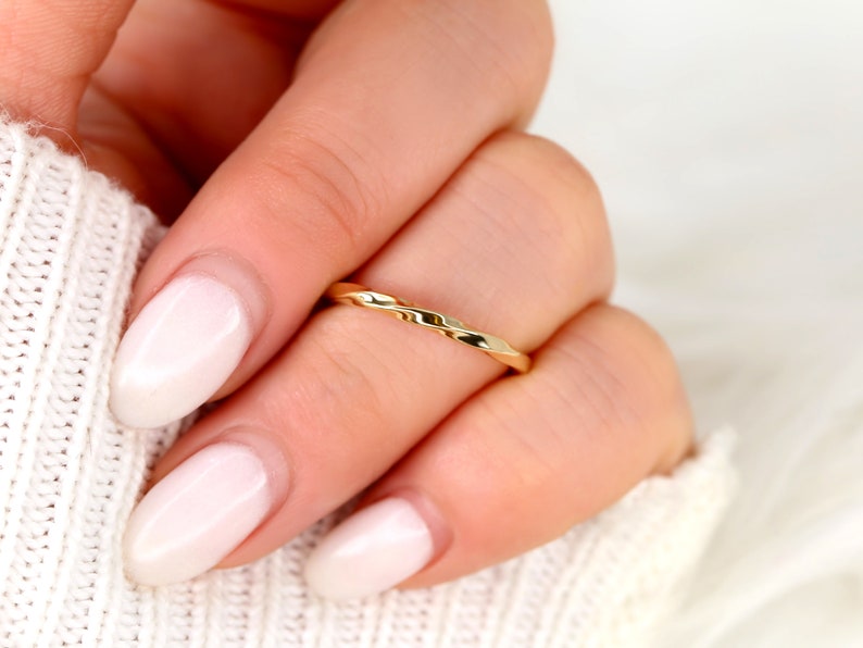 May include: A thin gold band ring with a hammered texture. The ring is worn on a finger with a light pink nail polish.