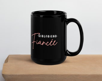 May include: Black ceramic mug with the word "Fiancée" in a pink script font, with the word "Girlfriend" crossed out above it. The mug has a large handle and sits on a wooden surface.