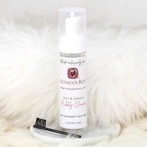 Può includere: Una bottiglia trasparente di "Gem & Jewelry Bubbly Cleaner" con un tappo a pompa bianco, una spazzola nera e una base bianca. La bottiglia ha un'etichetta con il testo "ROSADOS BOX" e "Bubbly Cleaner". La bottiglia contiene 75 ml di detergente.
