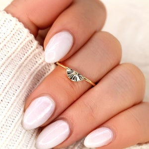 May include: A gold ring featuring a dainty fan-shaped design. The ring is worn on a finger with white nail polish.