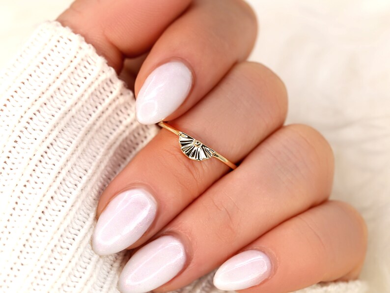 May include: A gold ring featuring a dainty fan-shaped design. The ring is worn on a finger with white nail polish.