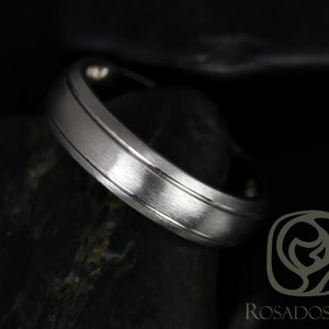 May include: A silver-colored ring with a brushed finish and a grooved design. The ring is set against a dark background, highlighting its metallic sheen. The Rosados Box logo is visible in the lower right corner.