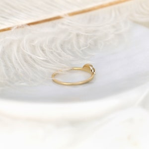 May include: A delicate gold ring with a small, clear gemstone set in the band. The ring is photographed against a white feather background.