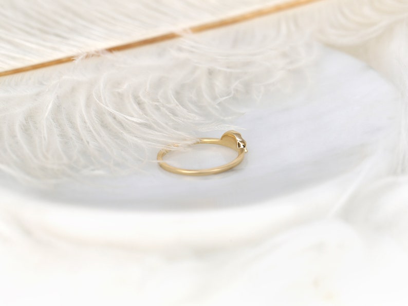 May include: A delicate gold ring with a small, clear gemstone set in the band. The ring is photographed against a white feather background.