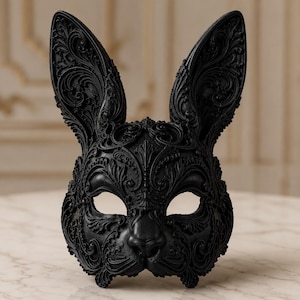 Può includere: Una maschera da coniglio nero opaco caratterizzata da intricati motivi a volute e filigrana in rilievo su tutta la superficie. La maschera presenta due orecchie alte e appuntite, aperture per gli occhi a mandorla e naso scolpito.