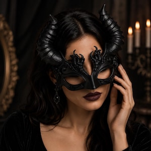 May include: A black masquerade mask featuring large, textured, curved horns extending from the sides and intricate, claw-like detailing around the eye openings. The mask has a matte finish and covers the upper portion of the face.