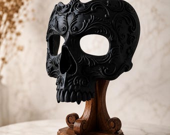 Pode incluir: Uma máscara de caveira preta fosca com padrões florais e de volutas em relevo intrincados na superfície. A máscara está montada num suporte de madeira castanho-escuro com um pilar curvo e uma base decorativa com pés em voluta.