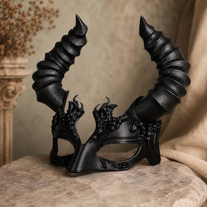May include: A black mask featuring two large, spiraled, textured horns extending upward. The mask has eye openings and intricate, raised decorative detailing along the brow and bridge of the nose, crafted with a matte finish throughout.
