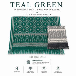 May include: Teal green Indonesian Troso handwoven fabric measuring 230cm x 115cm. The textile features a geometric diamond pattern in teal and grey with a fringed edge. A folded section displays a grey and white ikat-style pattern with fringe.