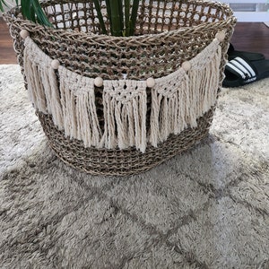 Könnte beinhalten: Ein geflochtener Korb aus Weide mit einer Makramee-Girlande mit Fransen, die um die Oberseite hängt. Die Girlande hat gleichmäßig verteilte Holzperlen.