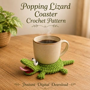 Op de afbeelding: Een groene gehaakte hagedis-onderzetter met witte ogen en een roze tong, die een beige keramische mok met donkere vloeistof ondersteunt. De tekst luidt Popping Lizard Coaster Crochet Pattern en Instant Digital Download.