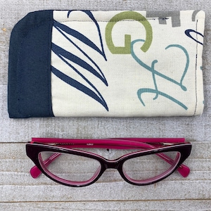 May include: A fabric glasses case with a navy blue accent and a cream-coloured patterned design. The case is paired with a pair of burgundy-coloured eyeglasses with pink accents.
