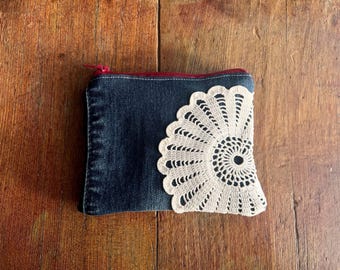 May include: A denim zipper pouch with a cream-colored, crocheted lace detail on the front. The pouch has a red zipper and a dark blue denim exterior. The lace is in a fan-like pattern.