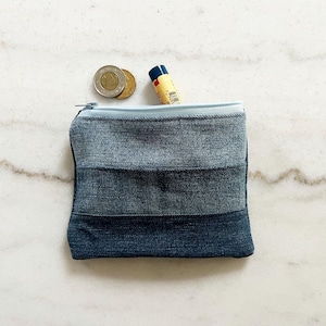 May include: A small, square denim coin purse with a light blue zip. The purse is made of three horizontal panels of varying shades of blue denim. Two coins and a tube of lip balm are visible inside the open purse.