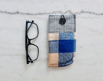 Könnte beinhalten: Eine Brille mit schwarzem Rahmen neben einer rechteckigen Stofftasche. Die Tasche hat einen oberen Bereich mit einem kleinen schwarz-weißen Karomuster und einen unteren Bereich mit einem blau-beigen Schottenkaro, verschlossen durch einen schwarzen Knopf.