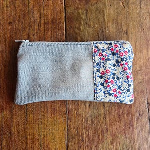 Peut inclure: Une pochette rectangulaire en jean avec une fermeture éclair. La pochette présente un panneau en tissu à motifs floraux avec des fleurs rouges et bleues sur un fond crème. Conçu pour les petits objets.