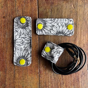 Peut inclure: Trois organiseurs de câbles en tissu avec un imprimé tournesol et des fermetures à bouton-pression jaunes. L'un est long et rectangulaire, un autre est plus court et rectangulaire, et le troisième est enroulé autour d'un câble noir. Sur une surface en bois.