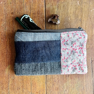 Könnte beinhalten: Eine rechteckige Reißverschlusstasche mit einem Patchwork-Design aus dunkelblauem Denim und einem hellgrauen Stoff mit kleinem rosa-grünem Blumenmuster. Ein schwarzer Stift und eine kleine braune Haarspange in Schildpattoptik liegen darauf.