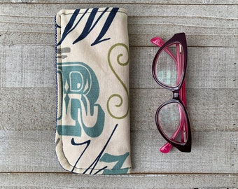 May include: A beige fabric glasses case with a blue, green, and white abstract pattern. The case is closed and lying on a wooden surface. Next to the case are a pair of pink-rimmed glasses.