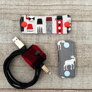 May include: Three fabric cable tidies in various patterns. One is red and black plaid, another has a pattern of black and white shapes with red accents, and the third is grey with white moose silhouettes. Each has snap closures.