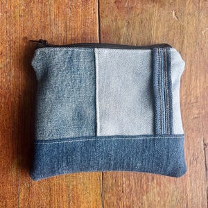 May include: A rectangular zipped pouch constructed from patchwork denim fabric. The design features three distinct shades of blue denim, including a dark blue base, a medium blue panel, and a lighter blue section, finished with a black zip fastener.