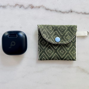 May include: A black square wireless earbud charging case with a logo next to a small square fabric pouch featuring a dark olive green diamond pattern, a light blue snap button closure, and a white side tab with a small metal ring attached.