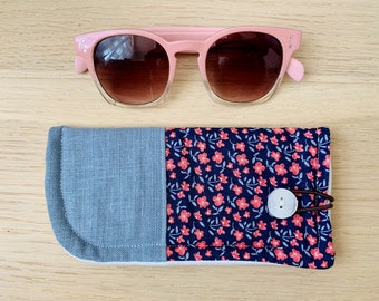 May include: A blue and pink floral fabric glasses case with a button closure and elastic strap. The case is closed and sitting on a wooden surface.