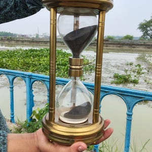 May include: An hourglass with a metallic gold finish frame and clear glass bulbs containing black sand. The structure features a circular top and base connected by four vertical pillars, with a central cylindrical connector between the two glass chambers.