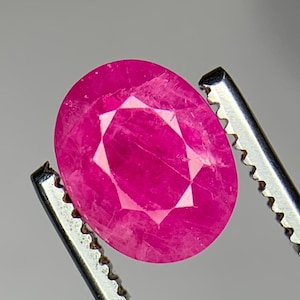 May include: A vibrant, oval-cut pink gemstone with a faceted surface, held by a pair of silver-toned metal tweezers. The stone displays a translucent quality with visible internal inclusions and a bright, saturated magenta-pink colour throughout.
